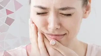 Kobieta z zamkniętymi oczami trzyma się za policzek i szczękę z bólu, co sugeruje, że cierpi na nadwrażliwość zębów lub ból zęba, powodujący dyskomfort w jamie ustnej.