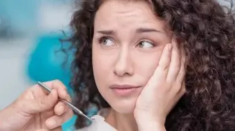 Kobieta z kręconymi włosami wygląda na zmartwioną i trzyma się za policzek, podczas gdy ręka dentysty, trzymająca narzędzie dentystyczne, znajduje się przed jej twarzą, co sugeruje, że może odczuwać ból zęba lub taki zębodół.