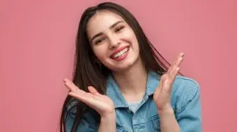 Młoda kobieta z długimi brązowymi włosami i jasnym uśmiechem, wzmocnionym przez licówki, podnosi obie ręce w pobliżu twarzy. Ma na sobie niebieską dżinsową koszulę i stoi na jednolitym różowym tle.