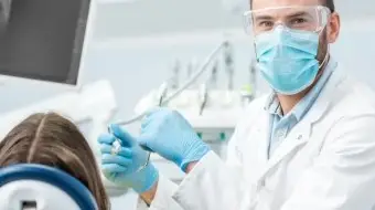 Dentysta w masce, osłonie twarzy i rękawiczkach przygotowuje się do leczenia pacjenta z odsłoniętymi szyjkami zębowymi siedzącego na fotelu dentystycznym, z widocznym sprzętem dentystycznym w tle.