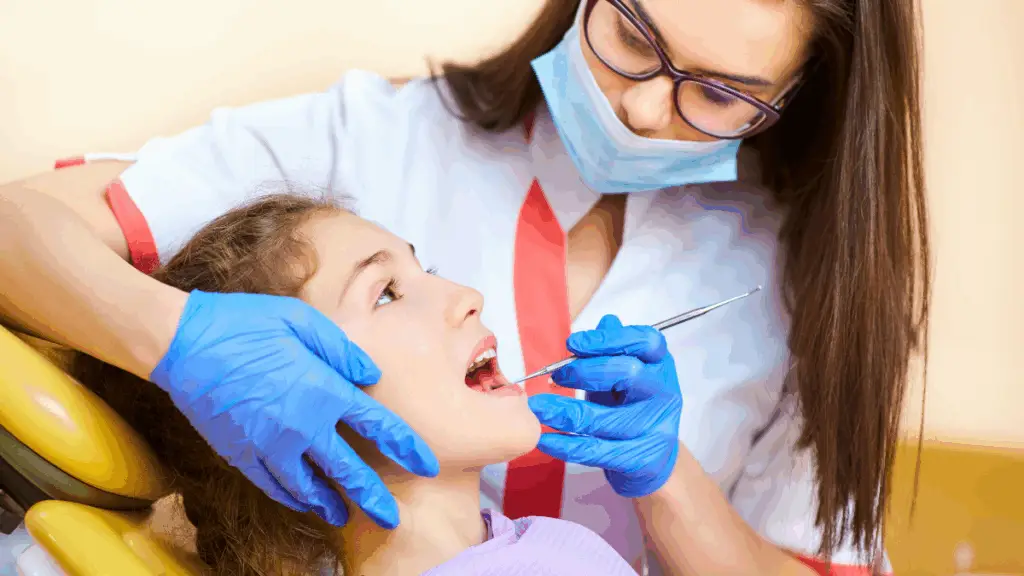 Dentysta w rękawiczkach, masce i okularach bada usta młodej dziewczyny siedzącej na fotelu dentystycznym, sprawdzając próchnicę u dziecka, gdy dziewczyna patrzy w górę, podczas gdy dentysta używa narzędzia dentystycznego.