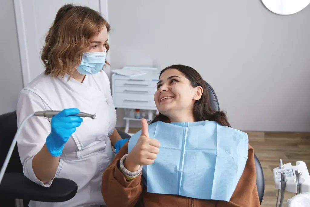 Uśmiechnięty dentysta w masce i rękawiczkach trzyma narzędzia dentystyczne gotowe do pomocy, podczas gdy pacjent na fotelu dentystycznym podnosi kciuk do góry, obaj wyglądają na szczęśliwych, gdy rozmawiają o płytce nazębnej i kamieniu nazębnym podczas wizyty.