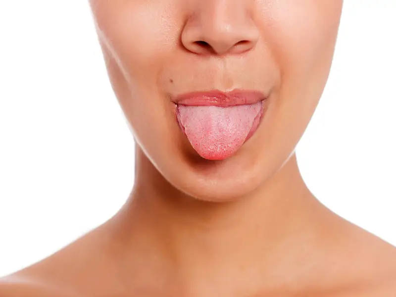 Osoba o jasnej karnacji wystawia język na białym tle. Widoczna jest dolna część twarzy i ramiona.