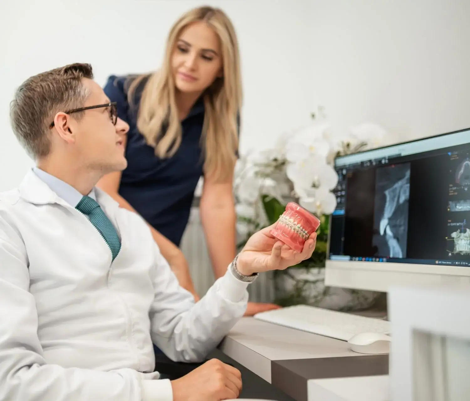 Stomatolog trzymający model zęba tłumaczy coś kobiecie, wskazując na zdjęcia rentgenowskie zębów wyświetlane na ekranie komputera w nowoczesnym gabinecie stomatologicznym.