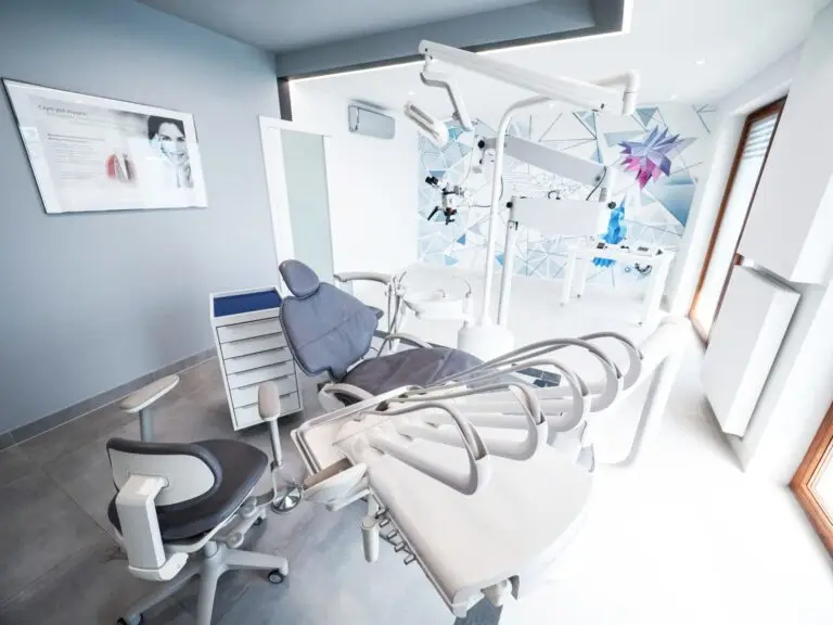 Nowoczesna klinika stomatologiczna z rozkładanym fotelem dentystycznym, sprzętem do endodoncji i narzędziami. Przestrzeń charakteryzuje się jasnym oświetleniem, szaro-białym wystrojem i geometryczną grafiką ścienną w tle.
