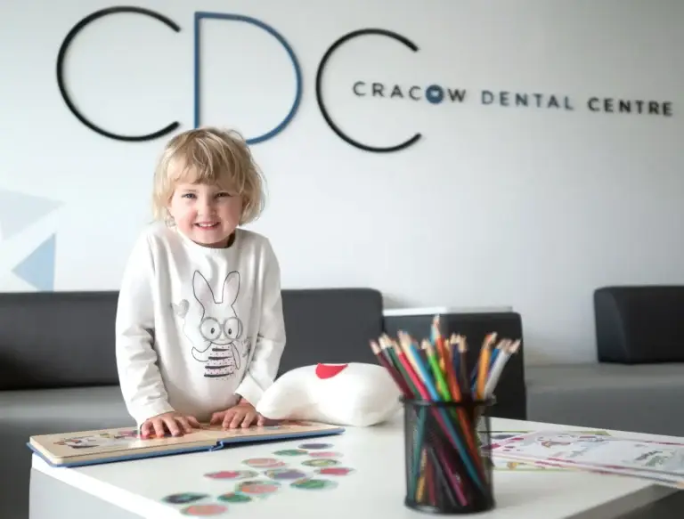Małe dziecko uśmiecha się, stojąc przy stoliku z kolorowymi kredkami i rysunkami w poczekalni, odzwierciedlając przyjazną atmosferę Pedodoncji w Krakowskim Centrum Stomatologii. Logo kliniki jest widoczne na ścianie z tyłu.