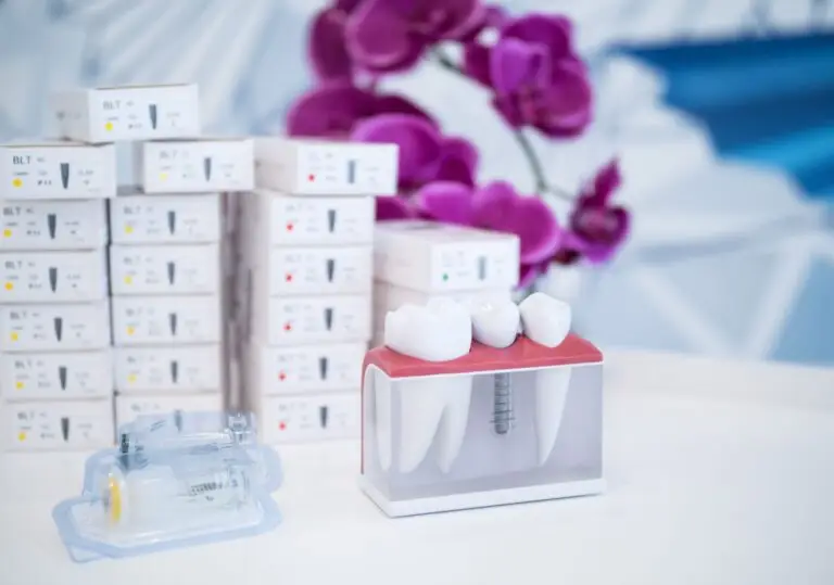 Model implantu dentystycznego ze sztucznymi zębami jest wyświetlany na stole, obok strzykawki i ułożonych w stos pudełek. Fioletowa orchidea zdobi rozmyte tło.