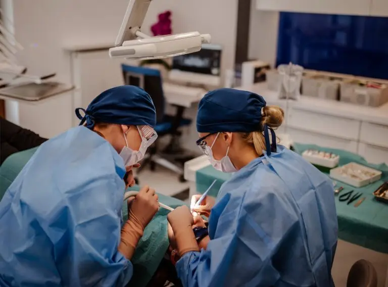 Dwóch dentystów specjalizujących się w stomatologii nosi niebieskie fartuchy chirurgiczne, rękawiczki, maski i czepki, wykonując zabieg dentystyczny na pacjencie w nowoczesnej klinice, z widocznymi w pobliżu narzędziami medycznymi i sprzętem implantologicznym.