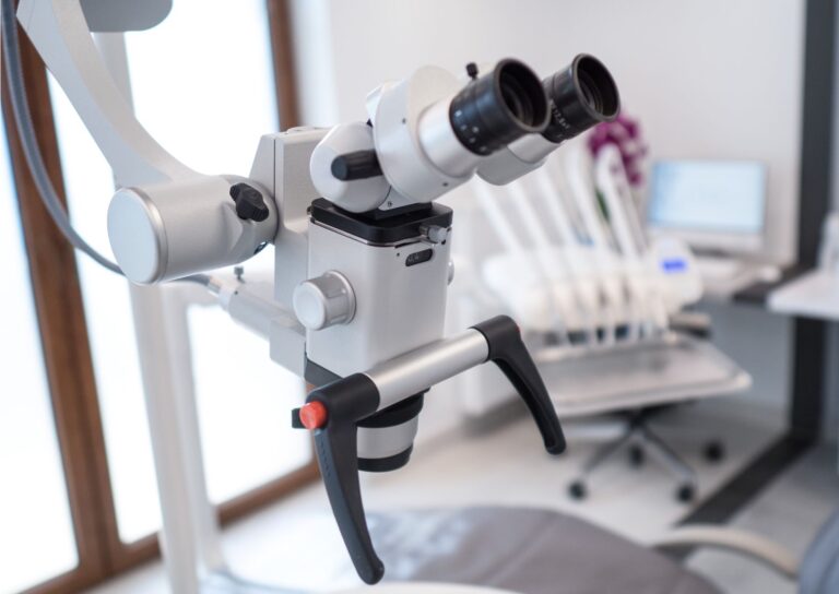 Zbliżenie mikroskopu stomatologicznego używanego w endodoncji w nowoczesnej klinice, z fotelem dentystycznym i komputerem rozmytym w tle. Sprzęt jest metalowy, z czarnymi okularami i uchwytami.