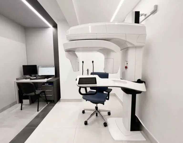 Nowoczesny gabinet stomatologiczny z panoramicznym aparatem RTG, niebieskim fotelem dla pacjenta i małym stanowiskiem z biurkiem i komputerem. Jasna, minimalistyczna przestrzeń jest idealna do zaawansowanych zabiegów, takich jak implantologia.