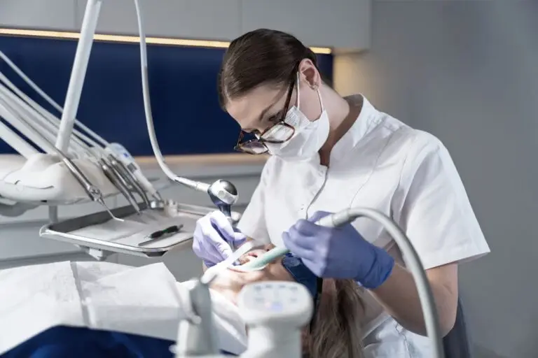 Stomatolog w białym fartuchu, masce i rękawiczkach wykonuje zabieg stomatologiczny na pacjencie leżącym w gabinecie stomatologicznym, używając narzędzi dentystycznych i jasnego światła do badania.